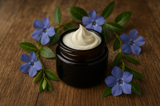 Stilfuldt billede af en lille, mørk glasbeholder med whipped tallow på en rustik træoverflade, omgivet af blå blomster og grønne blade, som symboliserer naturlighed og friskhed.